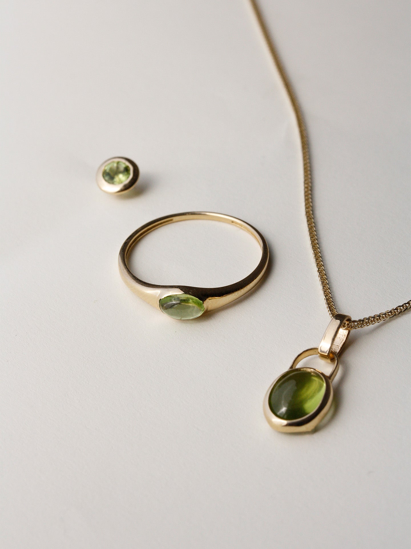 Flat lay of Padlock Charm Necklace pictured with the Mini Gem Signet and Peridot Bezel Stud on light grey background. 