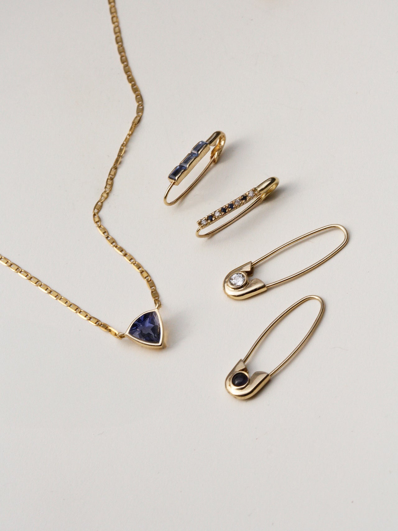 Flat lay of 14kt Yellow Gold Mini Diamond & Sapphire Safety Pin pictured with other seasonal styles + Iolite Trillion Necklace.