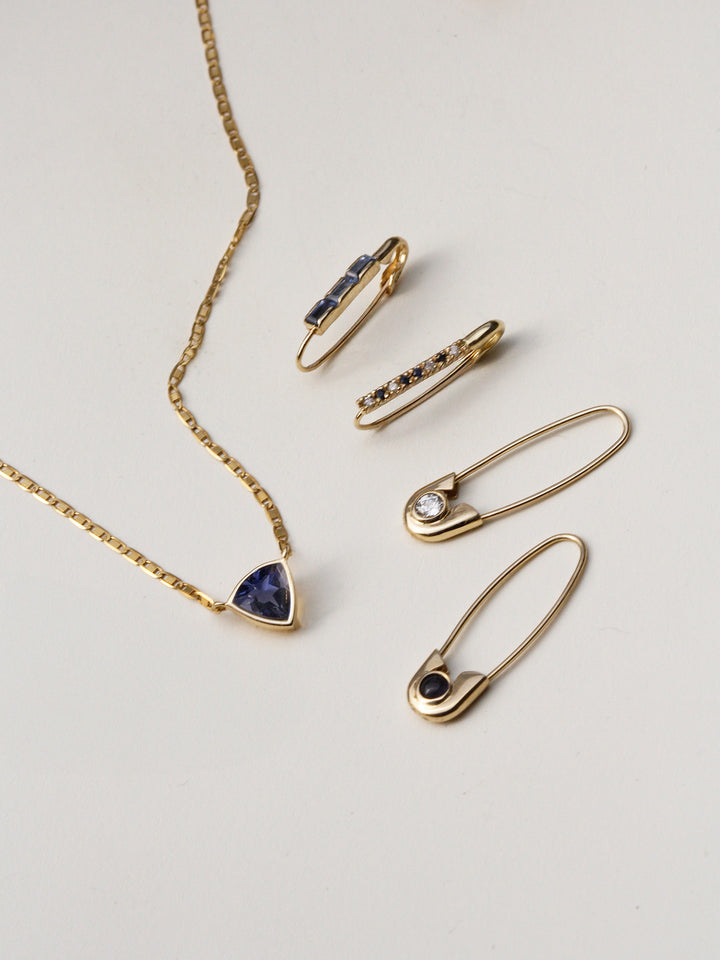 Flat lay of 14kt Yellow Gold Mini Diamond & Sapphire Safety Pin pictured with other seasonal styles + Iolite Trillion Necklace.